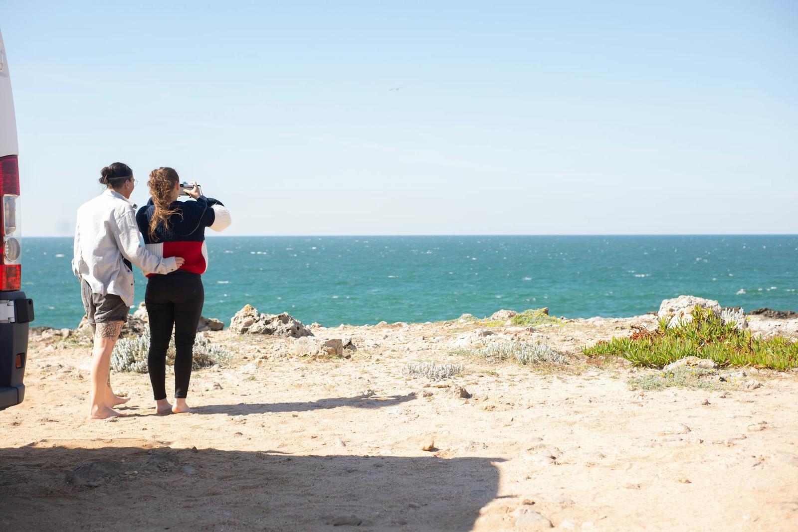 Two travellers standing beside their campervan on a clifftop, looking out to sea