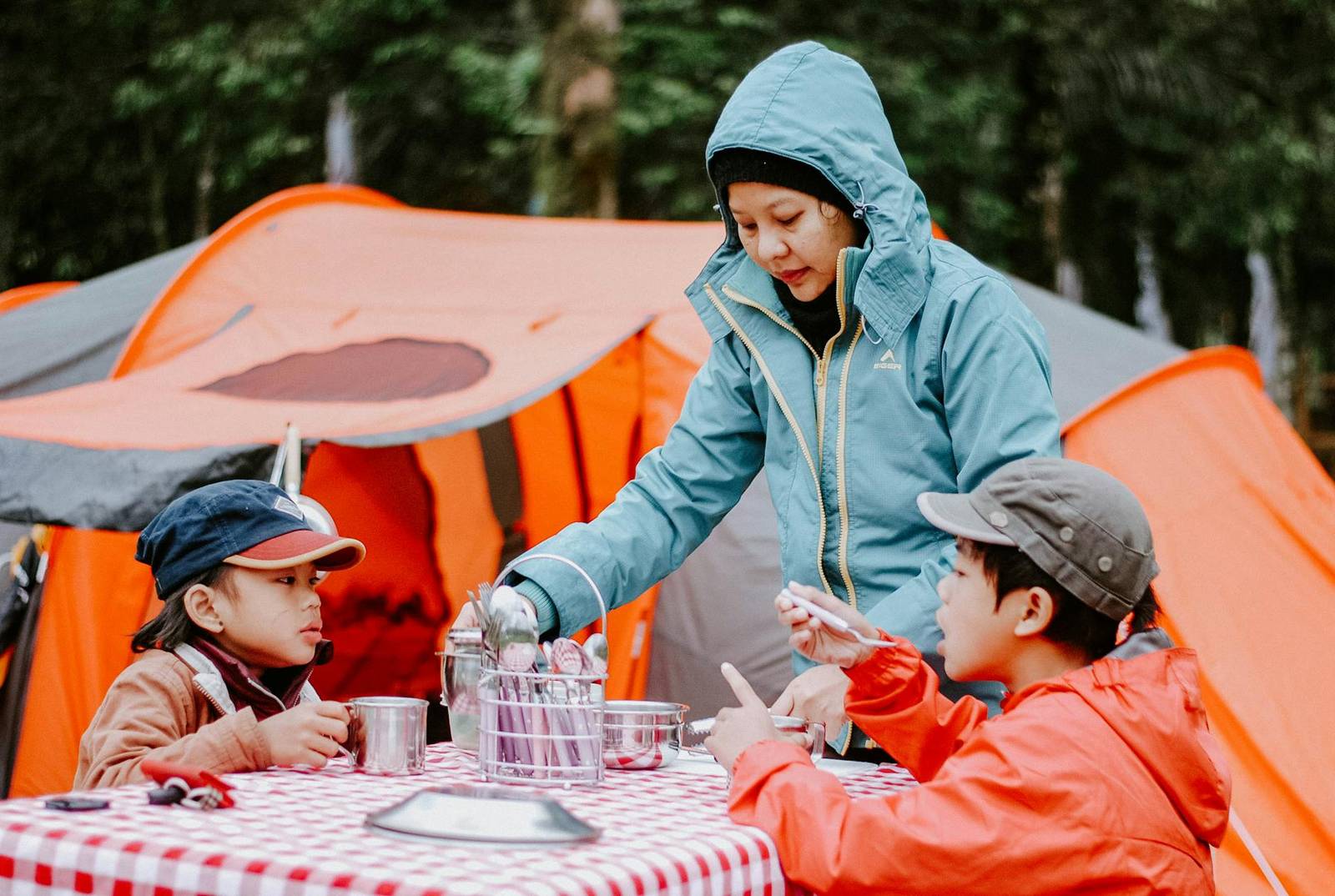 A mother serving two children at a camp table with a checked tablecloth, in front of orange tents