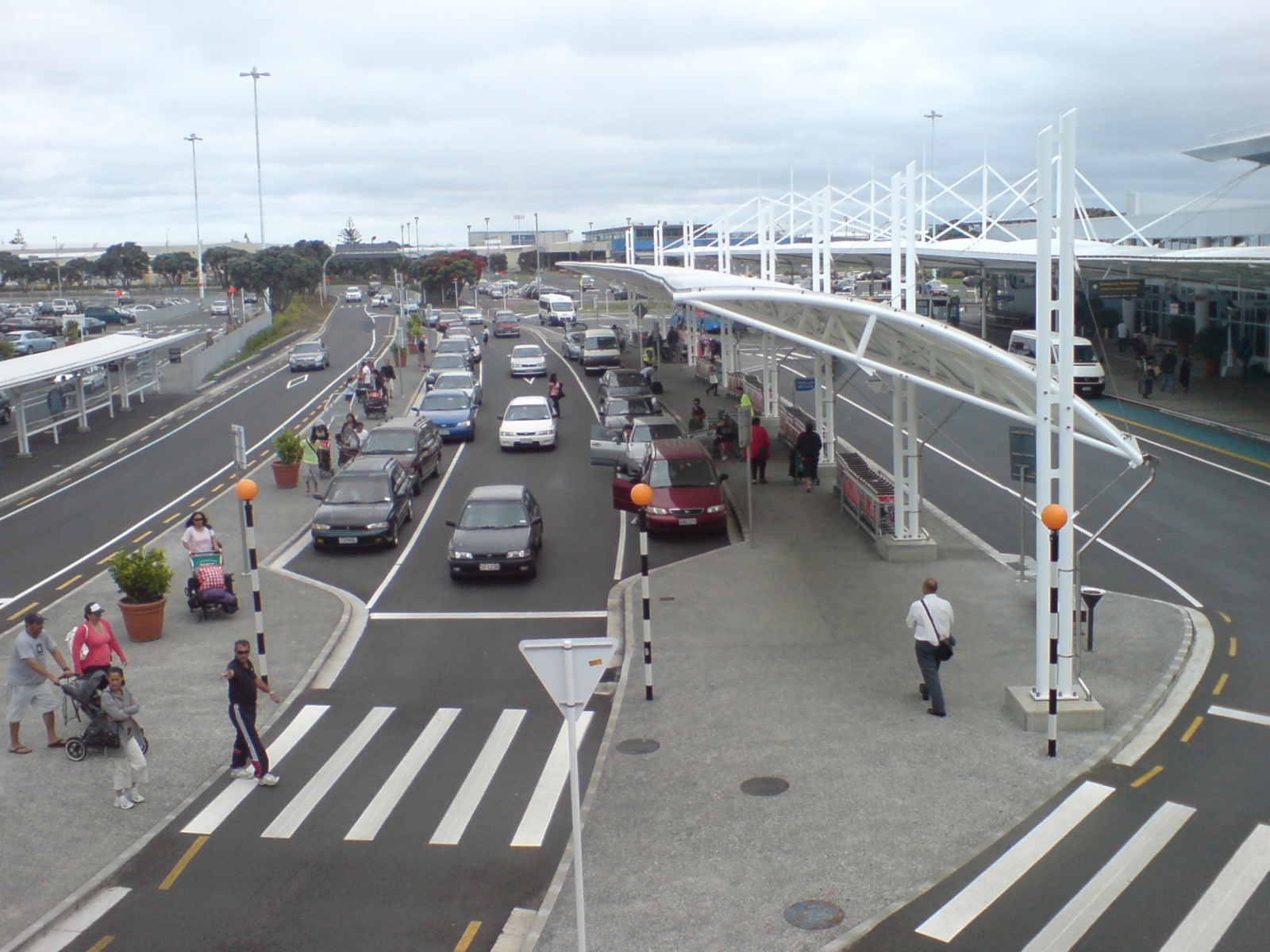 The kerbside forecourt outside Auckland Airport's international terminal, with cars queuing, covered walkways and luggage trolleys