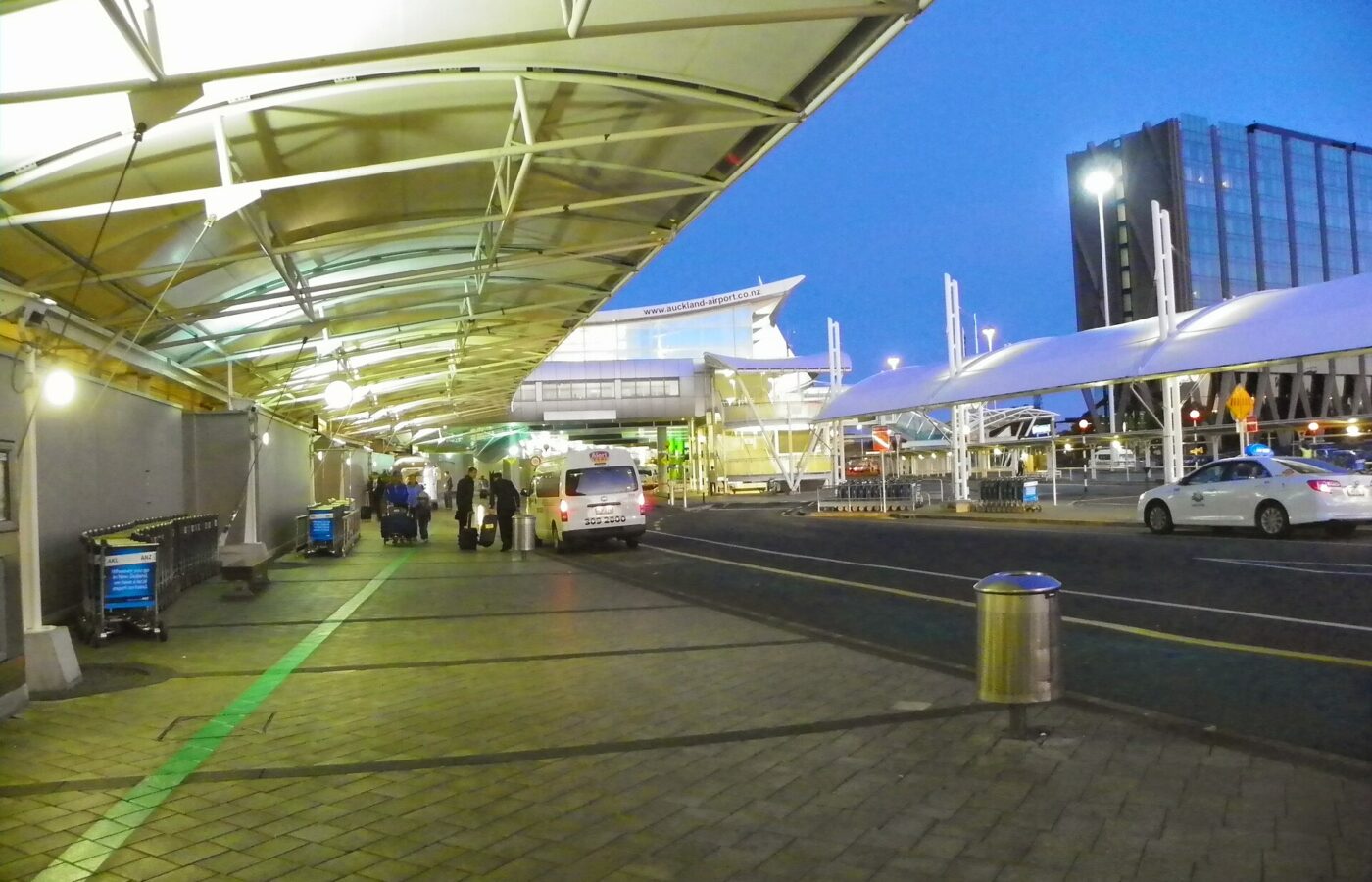 The covered forecourt outside Auckland Airport's international terminal at dusk, with taxis, luggage trolleys and the green walking line on the pavement