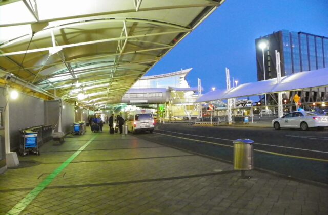 The covered forecourt outside Auckland Airport's international terminal at dusk, with taxis, luggage trolleys and the green walking line on the pavement