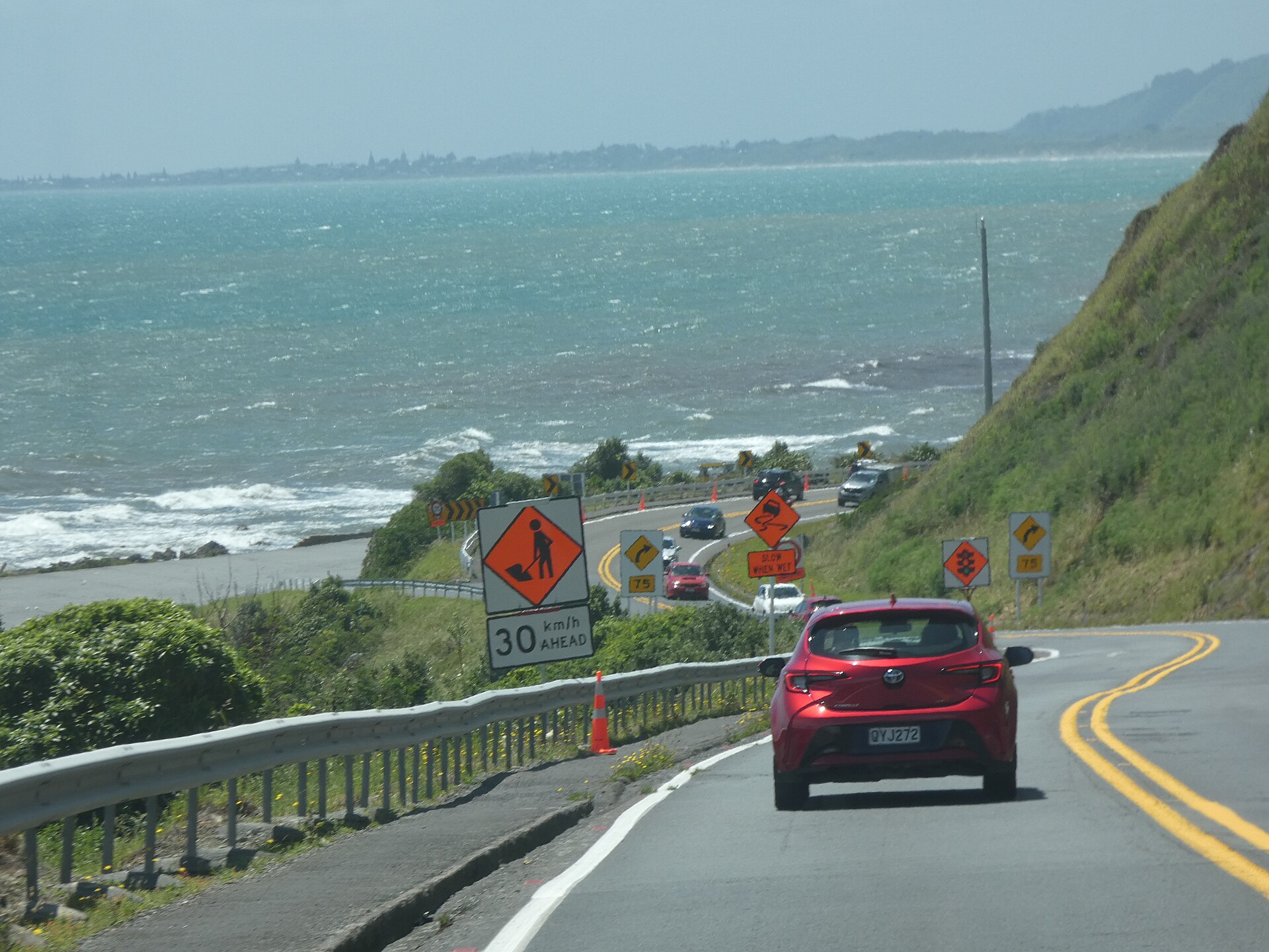 A New Zealand state highway curving along a coastline with roadworks warning signs and cars driving on the left