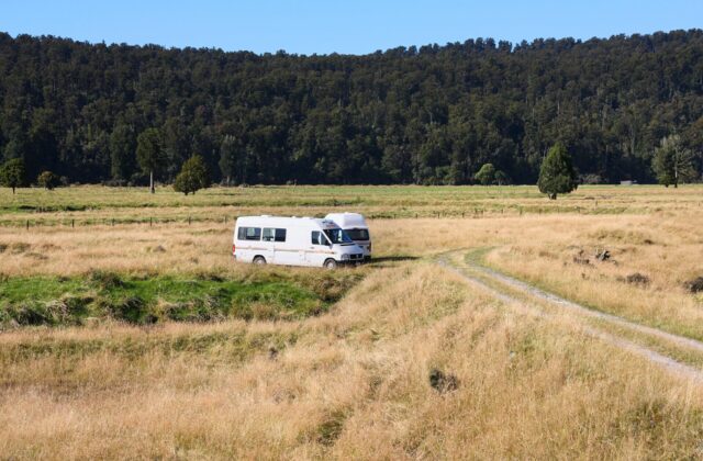 A white rental campervan parked on a grassy flat below native forest in New Zealand