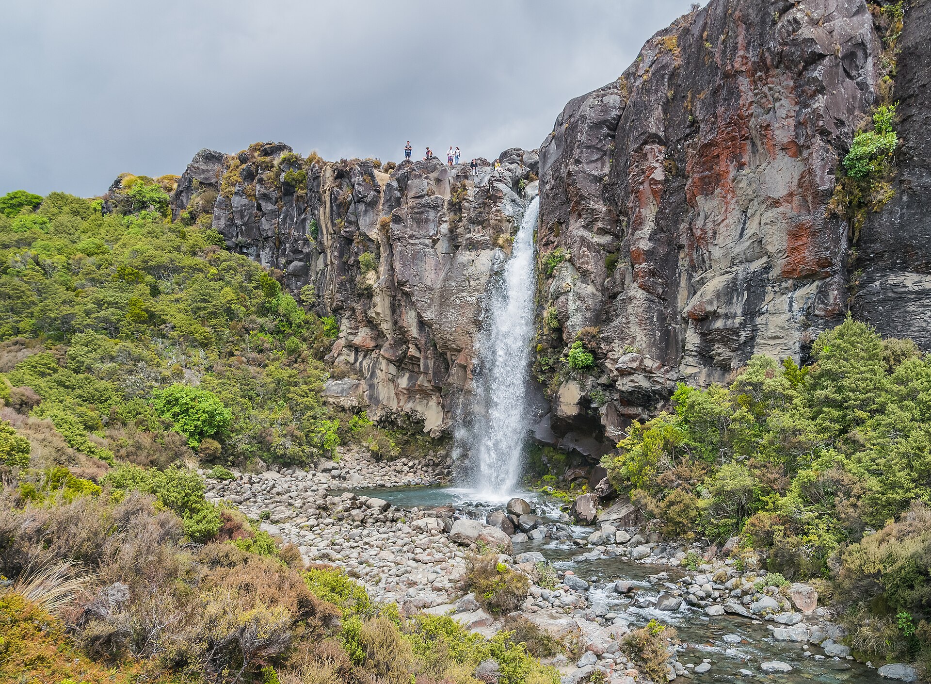 Taranaki Falls pouring over a dark lava cliff into a rocky pool surrounded by alpine scrub