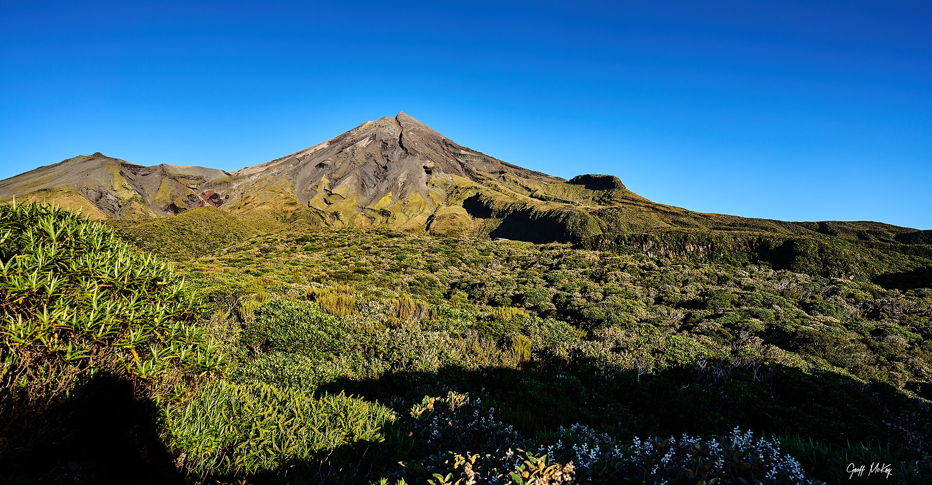 The snow-streaked cone of Taranaki Maunga rising above green alpine scrub under a clear blue sky